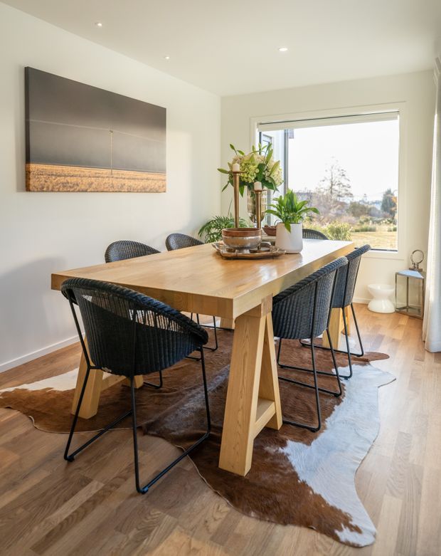 Bright contemporary dining room featuring a large solid timber dining table with angular trestle legs, dark woven dining chairs, light timber flooring, and a brown hide rug. A large abstract artwork hangs on the pale wall, with indoor plants and a broad window bringing in natural light and views of the landscape.