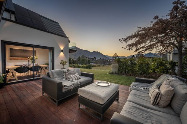 A dusk view of a contemporary home’s outdoor living deck, furnished with a large grey sectional sofa, matching armchair and ottoman around a low fire bowl. Dark timber decking, minimalist exterior walls, warm wall lighting and large glazed doors connect the deck to an illuminated dining area inside. A landscaped lawn and mountain backdrop are visible beyond.