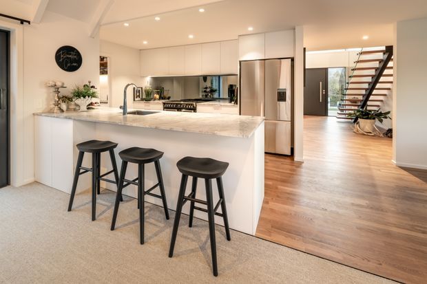 Bright open-plan kitchen with a large white island and waterfall-style stone benchtop, integrated white cabinetry, stainless-steel appliances, and three black bar stools. The kitchen opens to a timber-floored living or circulation area with a contemporary open staircase and dark-framed entry door. Recessed ceiling lighting and pale neutral finishes create a clean, welcoming interior.
