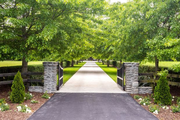 A formal landscaped entrance with a straight paved walkway leading through a tree-lined garden. The foreground features a dark asphalt approach, low timber fencing, stone gate pillars, black metal gates, clipped hedges, and neatly planted garden beds.