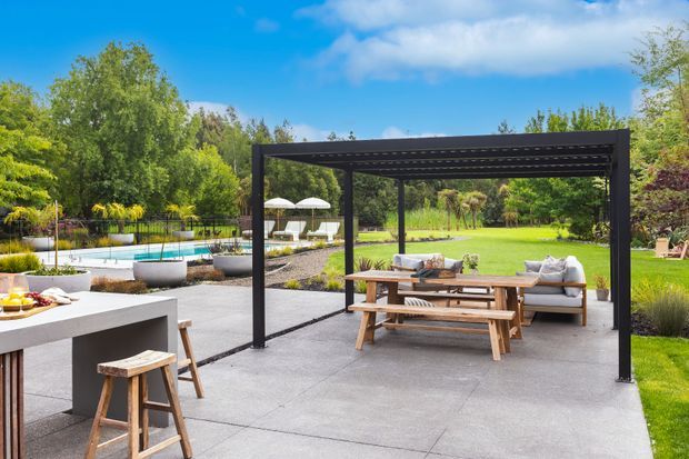 A spacious landscaped outdoor entertaining area with a dark timber pergola over a concrete patio. The patio includes a long wooden picnic-style dining table with benches, outdoor lounge seating, and a nearby outdoor kitchen or bar counter with stools. A swimming pool, umbrellas, lawn, mature planting, and garden beds are visible beyond.