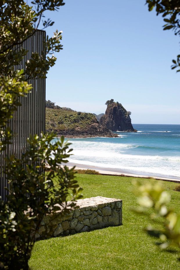 A landscaped coastal property overlooking a sandy beach and turquoise ocean, with a dark vertical-slat building facade partially screened by foliage at left. A low rough-stone retaining or garden wall sits in the manicured lawn in the foreground, while a rocky coastal headland forms the distant focal point.