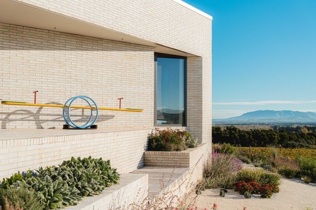 Exterior architectural photograph of a contemporary, rectilinear building clad in pale cream brick. A projecting upper volume creates a deep recessed balcony or wall zone, with a tall dark-framed window and built-in brick planter beds containing drought-tolerant landscaping. A yellow-and-blue recreational board or equipment rack is mounted to the wall. The property overlooks a broad rural or landscaped valley with distant mountains under a clear blue sky.