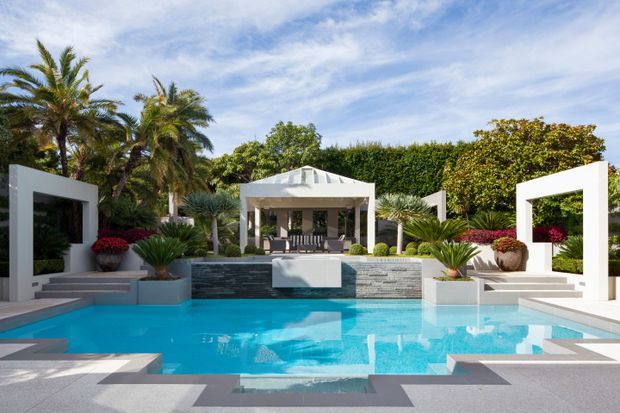 A bright, landscaped outdoor pool courtyard with a large turquoise swimming pool, pale stone paving, geometric white architectural pavilions, raised planters, tropical palms, clipped hedges, and a central covered outdoor seating pavilion with a hipped roof. The composition is symmetrical and resort-like, with strong modern geometric forms and lush planting.