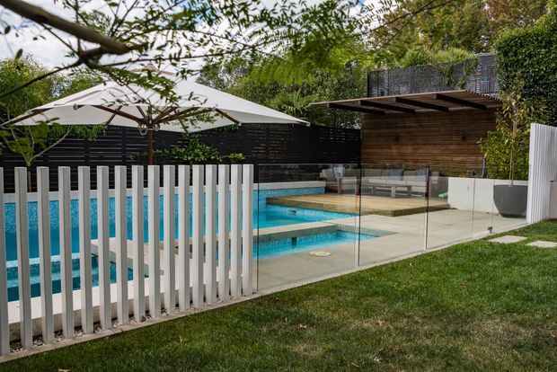 A landscaped backyard with a turquoise swimming pool enclosed by clear glass safety fencing and white vertical fencing. Large white umbrellas shade the poolside area, while a compact timber-clad pavilion or pool house with a flat projecting roof sits at the rear. Mature greenery and lawn surround the contemporary outdoor space.