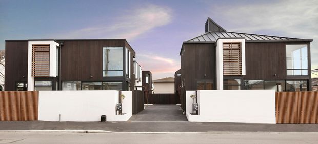 Street-facing view of a contemporary multi-unit residential development with two dark-clad, box-shaped dwellings. The buildings feature standing-seam metal roofs, large glazed openings, projecting upper-level volumes, and timber louvred window screens. White masonry boundary walls and vertical timber privacy screens frame the central driveway and pedestrian entry.