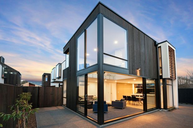 A contemporary multi-level residential house photographed from the exterior at dusk. The sharply angular volume features dark vertical timber-like cladding, extensive floor-to-ceiling glazing, black-framed windows and doors, and a projecting white side element with horizontal timber screening. Through the glazing, a warmly lit open-plan living and dining area is visible. The home sits within a fenced suburban setting with a paved outdoor area.