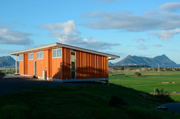 A compact single-storey contemporary cabin or small dwelling with vivid orange vertical timber cladding, a shallow mono-pitched roof, large rectangular windows, and elevated supports. It sits in an open rural landscape with expansive green fields and dramatic mountain peaks in the distance under a partly cloudy blue sky.