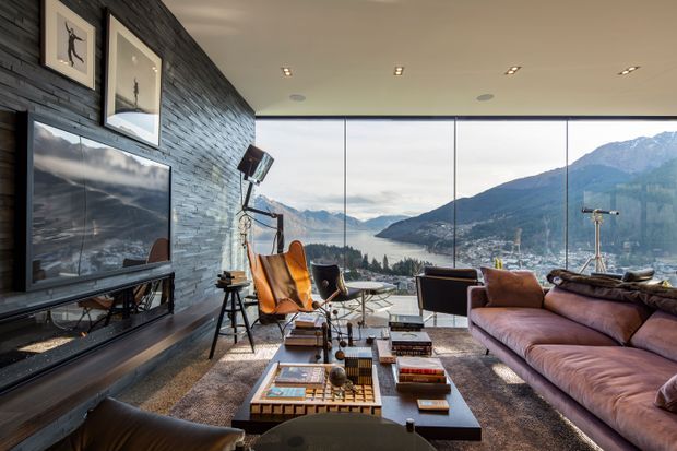 A sophisticated living room with expansive floor-to-ceiling glazing overlooking a mountainous lakeside landscape. The room features a dark stacked-stone feature wall with a wall-mounted television and framed artwork, a large brown sectional sofa, mid-century-style lounge chairs, a low timber-and-dark coffee table, layered rugs, and warm recessed lighting.
