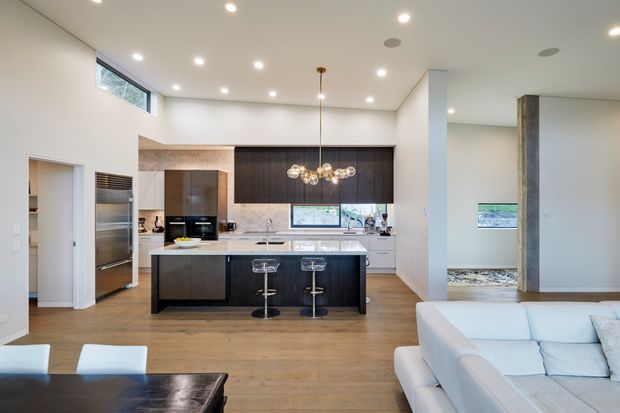 Open-plan contemporary kitchen and living area with a large dark timber island, white stone benchtops, integrated cabinetry, stainless-steel appliances, bar stools, timber flooring, and a white sectional sofa. Recessed ceiling lights and a sculptural pendant illuminate the bright, clean interior.