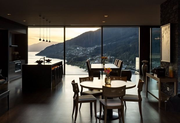 A sophisticated open-plan dining and kitchen interior with a round dining table and upholstered wooden chairs in the foreground, a dark kitchen island to the left, and full-height glazing overlooking a dramatic mountainous waterfront landscape. The room features dark stone wall cladding, dark cabinetry, pendant lights, polished dark flooring, and a warm, subdued atmosphere.