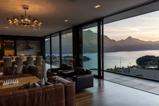 A sophisticated open-plan living and dining area with a dark, moody palette and expansive floor-to-ceiling glazing overlooking a mountain range and lake. The room includes large brown leather sofas, a timber coffee table, a dining table with upholstered chairs, dark kitchen cabinetry, timber flooring, recessed ceiling lights, and an ornate multi-arm chandelier. A broad terrace is visible beyond the glazing.
