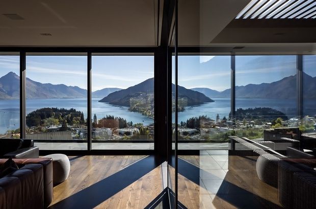 A modern interior living space with floor-to-ceiling glazing overlooking a dramatic mountainous lake landscape. The room has dark-framed glass walls, warm timber flooring, a dark leather sofa and contemporary woven lounge furniture. Strong sunlight creates long shadows across the floor, while a linear skylight or roof opening adds overhead daylight.
