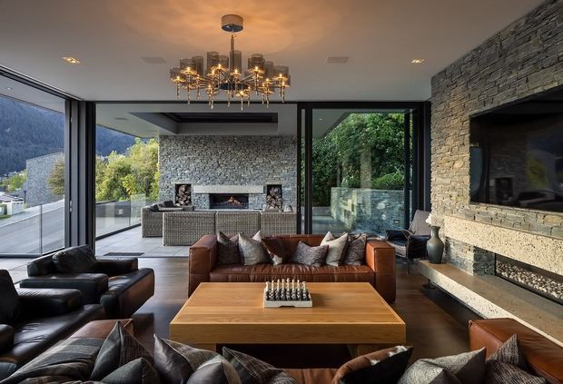 Luxurious contemporary living room with a large stone-clad feature wall and integrated fireplace, rich leather seating, a substantial timber coffee table, and an ornate chandelier. Full-height glazing opens the room to a terrace and landscaped mountain setting, while an outdoor fireplace is visible beyond.