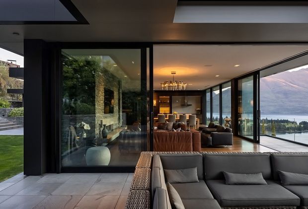 A sophisticated contemporary open-plan living and dining interior viewed from a covered outdoor threshold. The room features expansive black-framed floor-to-ceiling glazing, a large dark sectional sofa in the foreground, leather seating, a dining area and kitchen beyond, and strong visual connections to a lake and mountain landscape. Pale large-format tiled flooring, dark ceilings and joinery, and warm recessed and pendant lighting create a refined indoor-outdoor atmosphere.