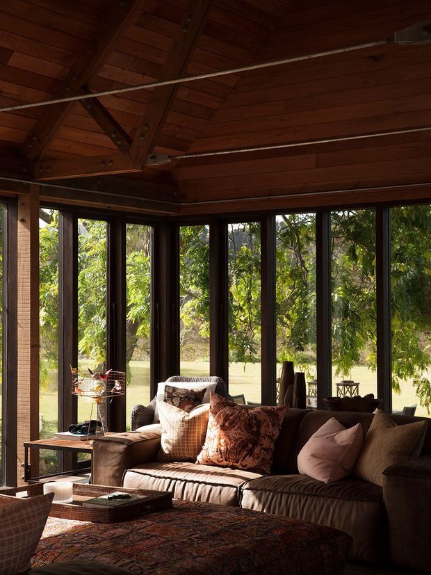 A warm, richly timber-lined living room with a vaulted exposed-beam ceiling and extensive dark-framed floor-to-ceiling glazing overlooking leafy grounds. A large brown sectional sofa, patterned cushions, a patterned ottoman, side tables, and a decorative pendant create a cosy, rustic interior.