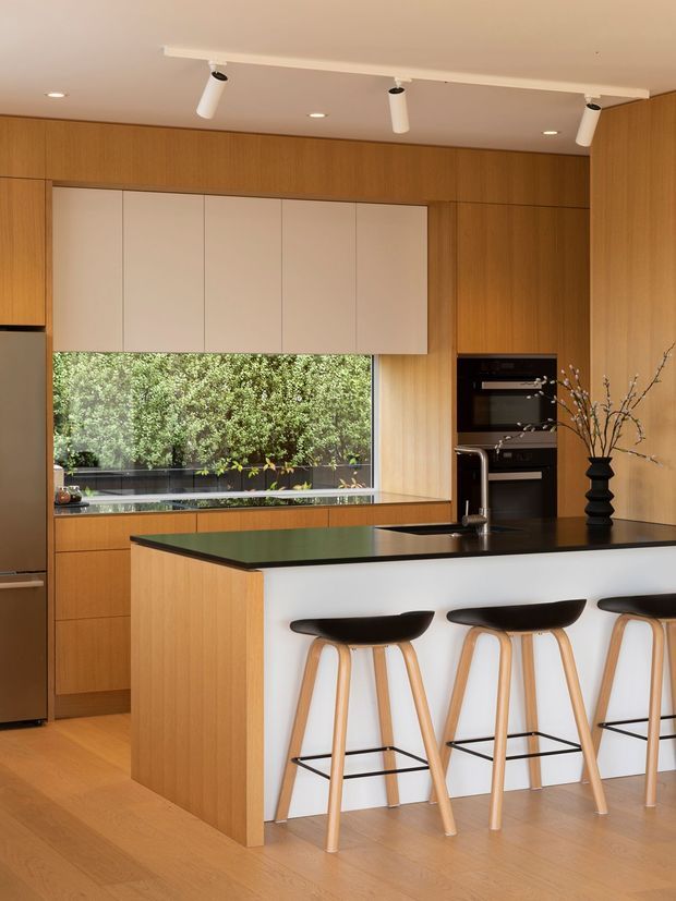 A warm contemporary kitchen with extensive light timber cabinetry, a large black island benchtop, white panelled overhead cupboards, integrated dark appliances, and three timber-and-black bar stools. A wide window overlooks greenery, while track and recessed lighting illuminate the clean, streamlined interior.