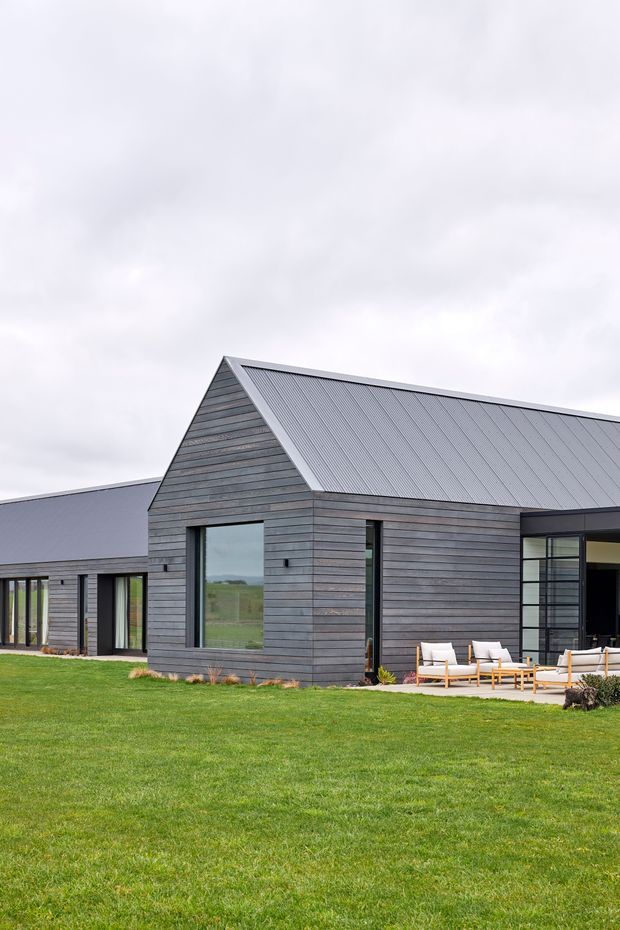 Exterior view of a contemporary single-storey rural residence with dark horizontal timber-look cladding, a prominent gabled corrugated metal roof, extensive black-framed glazing, and a covered outdoor seating area. The house sits within a broad manicured lawn under an overcast sky.