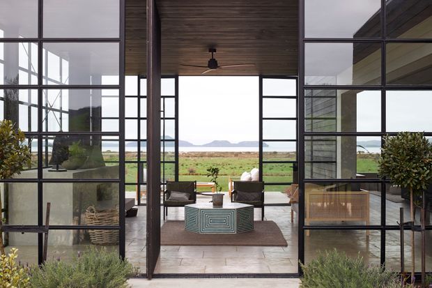 A sophisticated indoor-outdoor living area framed by expansive black steel-and-glass partitions. The space features a dark timber-lined ceiling, polished concrete or stone flooring, a central patterned coffee table, lounge chairs, potted greenery, and a broad rural landscape with water and hills visible beyond.