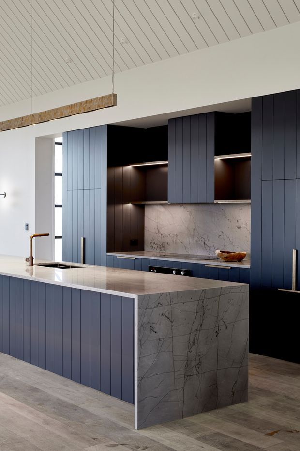 A refined contemporary kitchen featuring deep blue vertical-panel cabinetry, a large waterfall island clad in veined pale stone, matching stone splashback, dark open shelving with integrated lighting, and warm timber-look flooring. A white slatted ceiling and a linear pendant add architectural interest, while brass-toned tapware and hardware provide subtle accents.