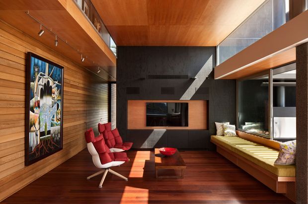 A warm, timber-lined contemporary living room with a rich dark-wood floor and ceiling, horizontal timber wall panelling, and a dramatic charcoal feature wall incorporating a recessed television niche. Red lounge chairs and a low timber coffee table sit opposite a built-in window seat with patterned cushions. Large glazing and strong natural sunlight create high-contrast shadows, while track lighting and integrated shelving complete the space.