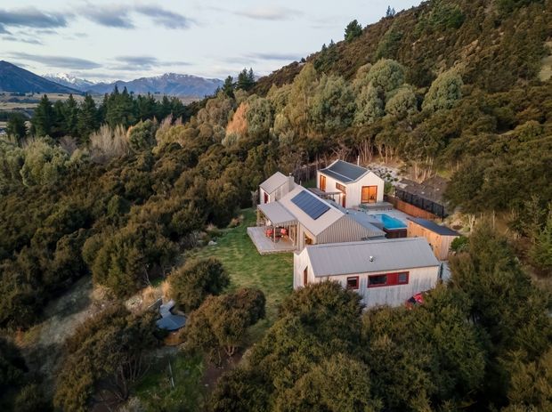 Aerial view of a secluded modern residential property nestled in dense bushland on a steep wooded hillside. The complex consists of several connected light-coloured buildings with pitched metal roofs, extensive glazing, and a small blue swimming pool, with distant mountain ranges visible beyond the site.