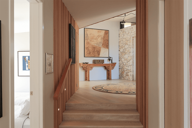 A warm, refined residential hallway viewed from the top of a short timber staircase. Vertical timber wall battens line both sides of the passage, with small recessed stair lights, pale timber flooring and steps, and a framed artwork above a traditional timber console table. A rugged light-coloured stone feature wall and round patterned rug anchor the landing, while an adjacent bedroom is partially visible through the doorway on the left.
