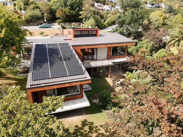 Aerial view of a contemporary multi-level home set within a densely planted suburban garden. The building has a low-pitched corrugated metal roof, extensive rooftop solar panels, warm timber-clad walls, large windows, and elevated sections supported above the landscaped site. Surrounding residences and mature trees are visible in the background.
