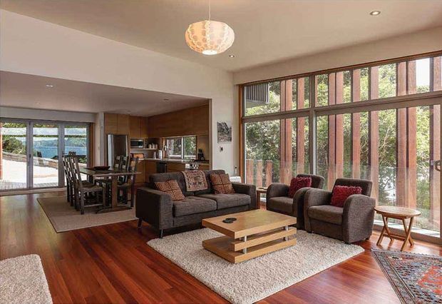 Open-plan living and dining area with a warm timber floor, large full-height glazed windows, a dark upholstered sofa and armchairs, and a low timber coffee table. A timber-lined kitchen and dining zone sit toward the rear, while the glazing looks onto leafy surroundings and an outdoor area. The room has recessed ceiling lights and a sculptural pendant, creating a relaxed, warm contemporary atmosphere.