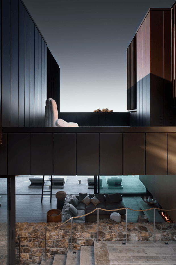 Dramatic architectural interior view of a multi-level contemporary residence, looking across a sunken living area toward dark vertically paneled volumes and an elevated courtyard or terrace. The space features a large sculptural lounge chair, grey sectional seating, a glass-topped or glass-sided feature area, exposed stone retaining walls, broad tiled steps, and a small fire feature. Strong geometric lines, deep shadows, and contrasting warm and cool lighting create a cinematic composition.