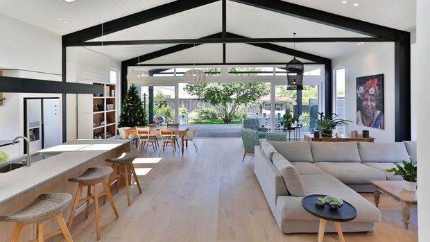 A bright, open-plan contemporary living space combining a kitchen, dining area and lounge. The room features pale timber flooring, white walls, extensive glazing, a large cream sectional sofa, a kitchen island with woven bar stools, and prominent exposed dark-painted structural beams supporting a high pitched ceiling. The space opens toward a landscaped outdoor area.