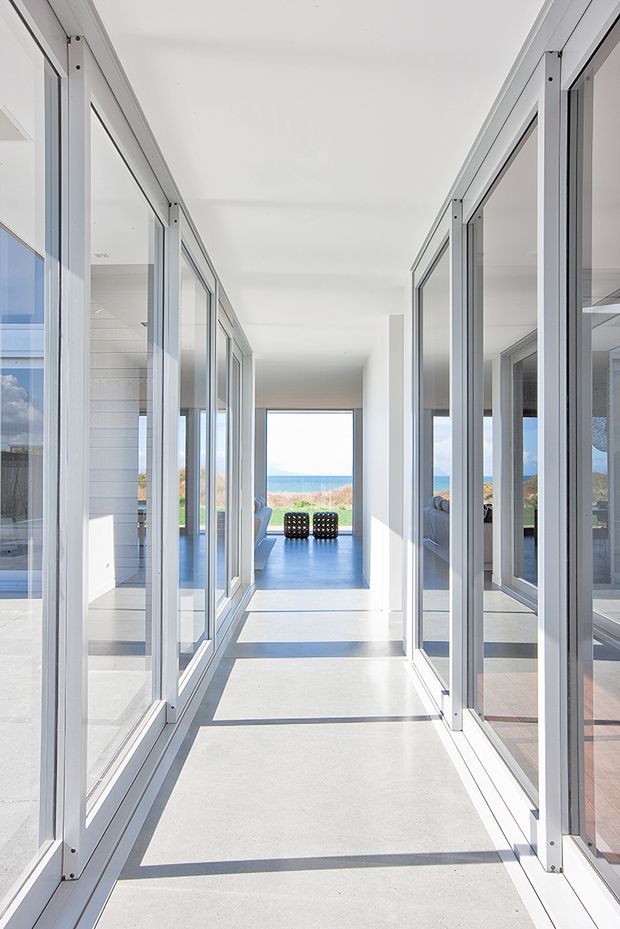 A bright, minimalist interior corridor lined on both sides with full-height glazed partitions and slim white frames. The polished pale floor reflects strong daylight, while the corridor opens toward a lounge and expansive ocean view beyond. Reflections reveal additional glazing and modern interior spaces, reinforcing the open, coastal architectural character.