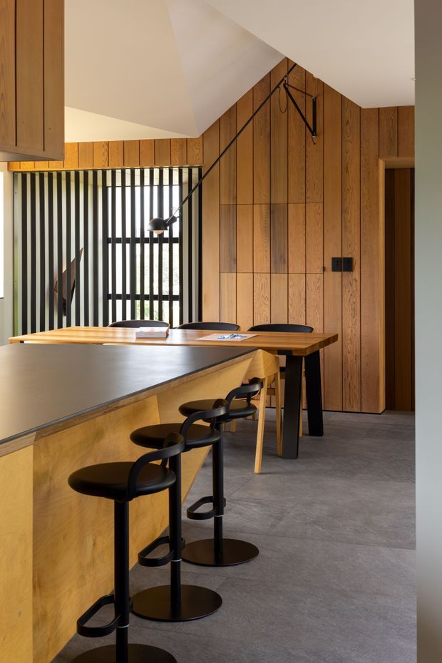 A contemporary kitchen and dining area featuring extensive warm timber wall panelling and cabinetry, a large dark benchtop island with multiple black bar stools, and a timber dining table. A tall black-and-white vertical slatted screen filters light near the rear, while a sculptural black articulated pendant lamp extends over the dining area. The room has a clean, refined material palette with grey flooring and a sloped white ceiling.
