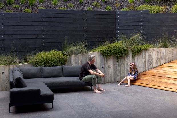 A landscaped outdoor entertaining area with a large dark sectional sofa on a broad grey-paved terrace. Concrete retaining walls with planted greenery separate the seating area from a raised garden bed and dark horizontal timber fencing. A warm timber deck extends to the right, creating a layered outdoor living space.