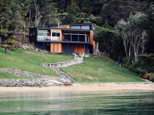 A modern dark-clad house set into a steep, heavily wooded hillside beside calm waterfront. The home features a compact rectilinear form, extensive glazing, projecting upper-level living spaces, warm vertical timber cladding, and stepped stone retaining walls and paths leading down to a sandy shoreline.
