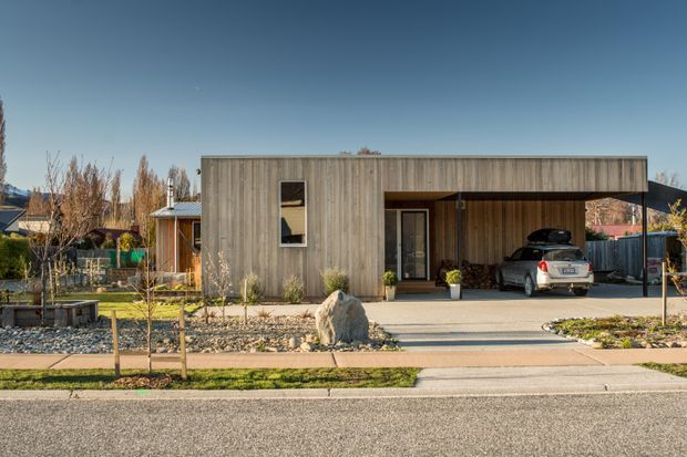 Single-storey contemporary house viewed from the street, with a long flat-roofed rectangular form clad in vertical weathered timber boards. A recessed entry and attached carport sit beneath the projecting roofline, while the front garden uses gravel, paving, young trees, and low planting. Residential surroundings and distant hills are visible in the background.