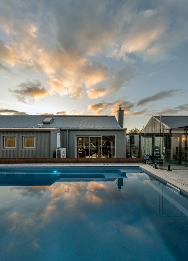 Exterior view of a contemporary single-storey residence with corrugated metal cladding and roofing, large glazed living areas, a timber pool deck, and a reflecting swimming pool in the foreground. The house is photographed at dusk beneath a dramatic cloud-filled sky.