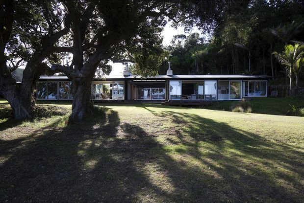 A low, single-storey modern house set within a leafy landscaped garden. The long horizontal facade has extensive floor-to-ceiling glazing, dark slender framing, pale wall surfaces, and a shallow low-pitched or flat roofline. A large established tree dominates the foreground, with tropical vegetation surrounding the property.
