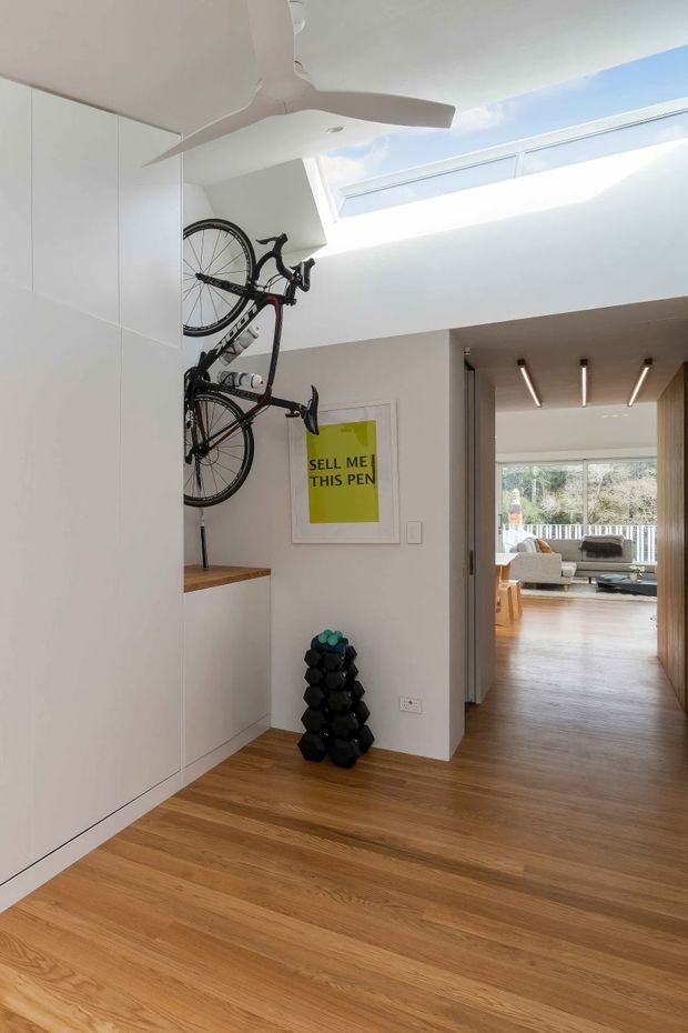 Bright contemporary interior hallway or entry area with warm timber flooring, full-height white built-in cabinetry, a wall-mounted bicycle rack, and stacked dumbbells. A large white ceiling fan and elongated skylight bring light into the space. Through the wide opening, a furnished living room with timber flooring and outdoor views is visible.