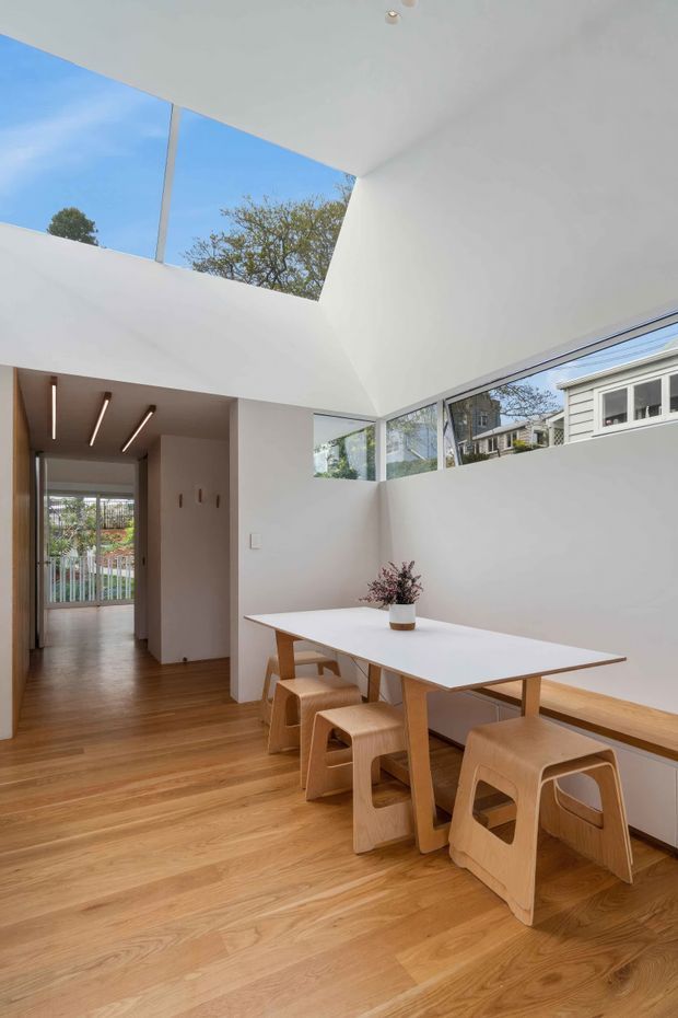 A bright, minimalist dining area with a long white dining table, pale timber stools, and a built-in timber bench. The room has warm timber flooring, white walls, high-level strip windows, and a dramatic skylight or roof opening that brings in abundant daylight. A hallway leads toward a glazed rear door, while neighbouring residential buildings and trees are visible through the windows.