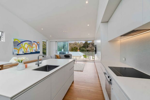 Bright contemporary open-plan kitchen with a large white island, integrated sink and dark cooktop, full-height white cabinetry, and a textured grey splashback. Timber flooring leads toward a glazed living area and balcony beyond, with colourful wall art and restrained modern furnishings.