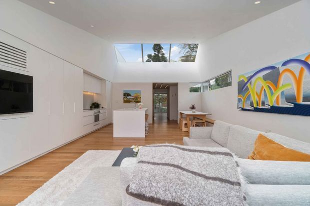 Bright open-plan contemporary living space with a white built-in media and storage wall, compact kitchen island, timber flooring, dining area, and a pale sectional sofa. A large skylight and high ceiling bring in natural light, while colourful abstract wall art adds a vivid accent.