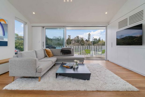 Bright contemporary living room with a large light-grey sectional sofa, black low-profile coffee table, pale textured rug, and warm timber flooring. Full-height sliding glass doors open to a balcony with white railings and greenery beyond. A white built-in media wall with horizontal ventilation grilles and wall-mounted television occupies the right side.