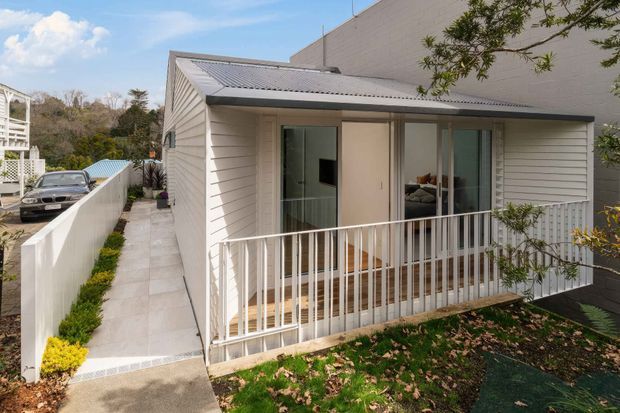 Exterior view of a compact, single-storey dwelling with a light weatherboard facade, corrugated metal gable roof, covered front balcony, sliding glazed doors, and white metal balustrading. A paved side walkway and landscaped garden border the home, with a taller adjoining wall visible behind it.