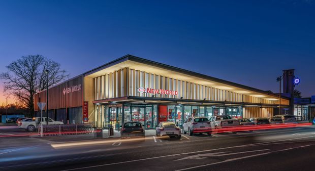 A dusk exterior view of a New World supermarket in a contemporary retail building. The prominent street-facing facade combines illuminated vertical fins, extensive glazed storefronts, and darker metal-clad wall sections, with a covered entry and surrounding car park visible in the foreground.