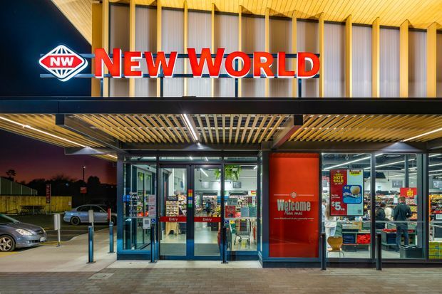 Exterior evening view of a New World supermarket entrance, featuring prominent red signage, illuminated glazed automatic doors, a covered forecourt with timber-look slatted soffits, and visible retail interiors beyond the glazing.