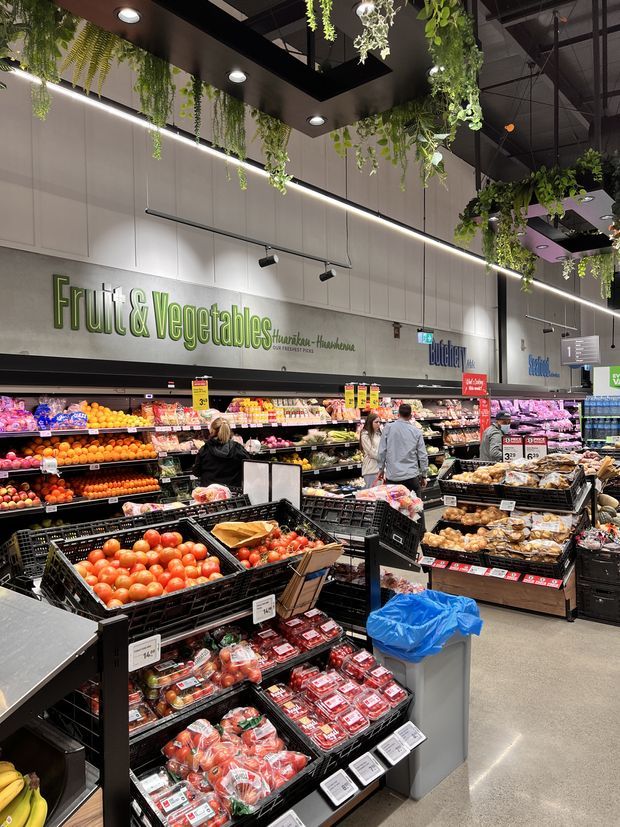 A brightly lit supermarket produce department with extensive displays of fruits and vegetables in black crates and refrigerated shelving. The space features prominent “Fruit & Vegetable” signage, high ceilings with exposed services, linear lighting, and abundant hanging greenery used as decorative overhead planting.