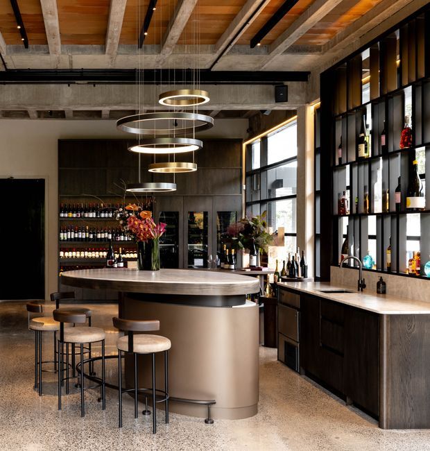 A sophisticated contemporary home bar or hospitality-style entertaining space with a large curved central island, four upholstered bar stools, extensive dark built-in cabinetry and open bottle shelving. The room features a polished light stone benchtop, integrated sink, wine and spirits storage, exposed concrete beams, warm timber ceiling panels, black-framed glazing, and suspended circular ring pendant lights. The palette is warm and refined, combining dark timber, concrete, brass-toned lighting, and neutral stone surfaces.