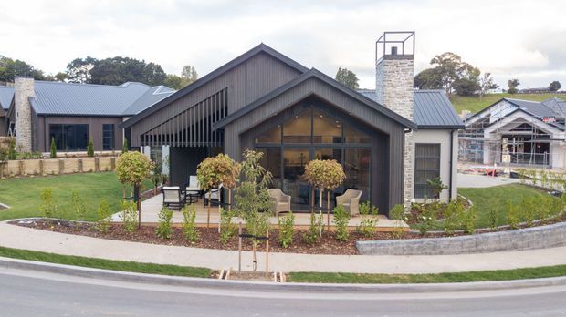 Exterior view of a contemporary, single-storey gabled home with dark vertical cladding, grey metal roofing, large glazed gable frontage, and a prominent stone chimney. The landscaped front garden includes a paved terrace, young planting, retaining edging, and a curved pedestrian path. Other homes and a partially constructed building are visible in the background.