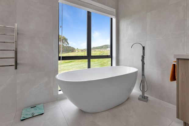 Bright contemporary bathroom featuring a freestanding white oval bathtub positioned beside a large black-framed window overlooking a rural landscape. The room has large light-grey wall and floor tiles, a sculptural floor-mounted bath filler, a stainless-steel towel rail, and light timber vanity cabinetry.